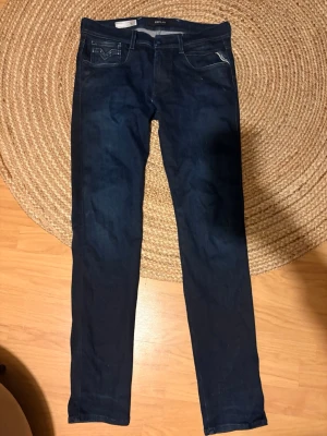 Mörkblå slim fit jeans från Replay - Säljer ett par mörkblå slim fit jeans från Replay med klassisk femficksdesign och snygga detaljer på bakfickorna. Jeansen har raka ben och är tillverkade i ett stretchigt material som ger en skön passform. Storlek 33