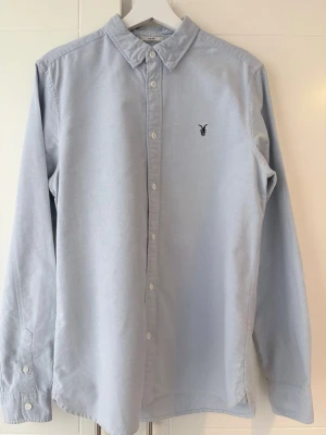 Ljusblå skjorta från AllSaints Spitalfields - Säljer en ljusblå skjorta från AllSaints Spitalfields med klassisk krage och knappar framtill. Skjortan har ett diskret broderat logomärke på bröstet och är långärmad med knappslå vid ärmslut. Perfekt för dig som gillar stilrena plagg.