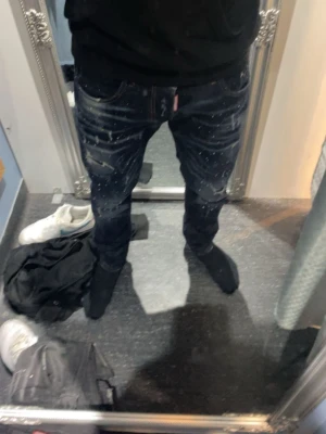 Mörkblå DSQUARED2 jeans med slitningar - Säljer ett par mörkblå jeans från DSQUARED2 med coola slitningar och färgstänk på både fram- och baksida. Jeansen har klassisk femficksmodell, raka ben och en patch med DSQ2-logga bak i midjan. Perfekta för dig som gillar streetwear och en edgy stil.