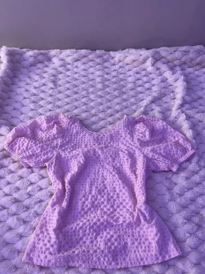 Ljuslila blus med puffärm - Söt ljuslila blus med puffiga korta ärmar och rund halsringning. Blusen har ett strukturerat tyg med små prickar som ger en trendig look. Perfekt att matcha med jeans eller kjol för en fräsch stil.