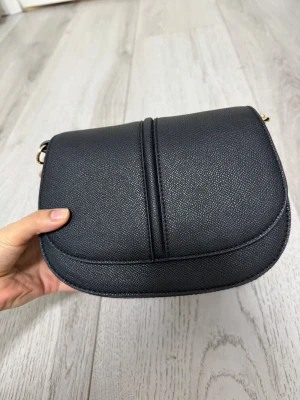 Svart axelväska i konstläder - Snygg svart axelväska i konstläder med rundad form och guldfärgade detaljer. Väskan har ett stilrent och minimalistiskt utseende med en dekorativ söm på locket. Perfekt storlek för det viktigaste och enkel att matcha till olika outfits.