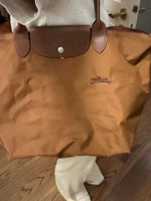 Brun Longchamp Le Pliage handväska - Säljer en klassisk brun Longchamp Le Pliage handväska i nylon med detaljer och handtag i brunt läder. Väskan har en enkel knappstängning och broderad logga på framsidan. Perfekt storlek för att bära med sig allt man behöver. Använd 1 gång❤️