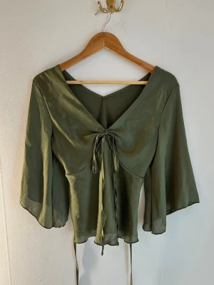 Grön topp - Snygg olivgrön omlottopp med v-ringning och vida ärmar. Toppen har knytband både fram och bak för en justerbar passform. Tillverkad i ett tunt, ”satin” lätt material som ger ett flowy intryck. Perfekt för dig som gillar boho-vibbar och vill ha något unikt i garderoben.