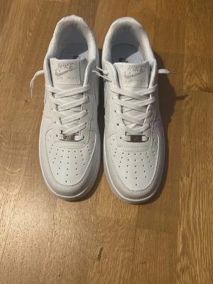 Nike Air Force 1 vita sneakers - Klassiska helvita Nike Air Force 1 sneakers i lågt utförande. Skorna har en stilren design med perforerad tå, vita skosnören och Nike-logga på sidan. Tillverkade i slätt läder med gummisula för grym komfort och streetstyle. Passar 43/44 och är helt oanvända