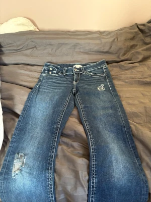 Blå slitna jeans från Gina Tricot - Ett par blå jeans från Gina Tricot med slitna detaljer på låret och vid fickan. Klassisk femficksmodell med normal midja och raka ben. Jeansen har vita kontrastsömmar och är tillverkade i denim med en snygg tvätt.