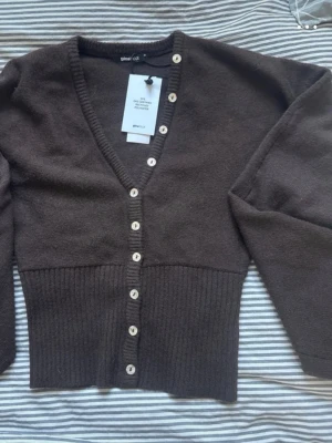 Brun kofta från Gina Tricot XS - Mysig brun kofta från Gina Tricot med v-ringning och pärlemorknappar framtill. Modellen är croppad med bred ribbad midja och långa ärmar. Tillverkad i mjukt material som passar perfekt till lager på lager-stil.