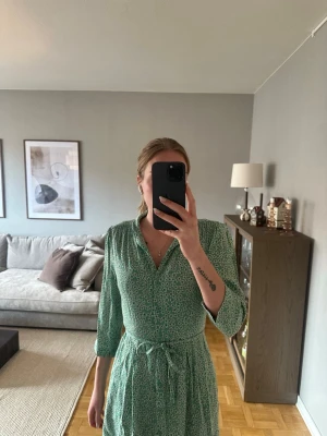Grön blommig kortklänning med knyt - Snygg grön kortklänning med småblommigt mönster och knytband i midjan. Klänningen har trekvartsärm och v-ringad hals. Från Lindex (Holly & Whyte) andvänd ett fåtal gånger. 