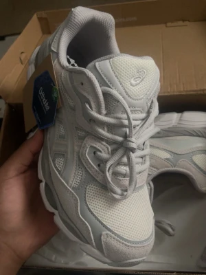 Vita Asics sportskor med mesh och mocka - Säljer ett par vita Asics sportskor med ljusgrå detaljer. Skorna har meshpaneler för bra ventilation, mockaliknande partier och klassisk snörning. Ortholite-innersula för extra komfort och en chunky sula som ger en modern och sportig känsla.