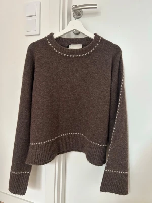 Brun stickad tröja från Neo Noir - Brun stickad tröja från Neo Noir  med vita detaljer längs halsringning, ärmar och nederkant. Tröjan har en rund hals och långa ärmar med breda ribbade muddar. Använd enstaka gånger, nypris 899kr