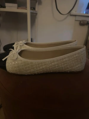 Vita ballerinaskor med lackad tå - Stilrena ballerinaskor i vitt tyg med struktur och glittriga detaljer. Skorna har rundad tå i svart lack och en söt rosett framtill. Klassisk ballerinaform med låg klack och mjuk innersula för extra komfort.