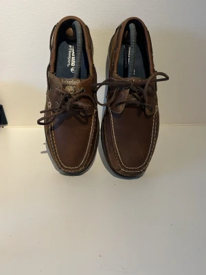 Timberland earthkeepers seglarskor - Ett par bruna seglarskor med vita kontrastsömmar i storlek 40. Skorna är knappt använda, utan har mest stått i skohyllan. Sulorna har inga märken, utom mild smuts. Riktigt snygga skor inför sommaren! Tveka inte att ställa frågor! 