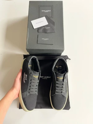 Svarta Saint Laurent sneakers - Snygga svarta sneakers från Saint Laurent med vit logga på sidan och guldfärgad text på plösen. Skorna har en klassisk låg siluett, svarta skosnören och vit sula. Tillverkade i canvas och läderdetaljer för en stilren look. Skickas samma dag dem köps, skriv gärna vid frågor😊