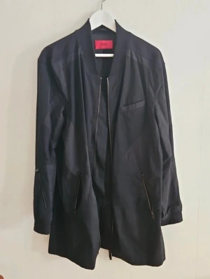 Svart bomberrock från Hugo Boss - Snygg svart bomberjacka från Hugo med längre passform och dragkedja framtill. Jackan har ribbade muddar vid kragen, två snedställda fickor med dragkedja och en bröstficka. Tillverkad i ett slätt material som ger en stilren look.