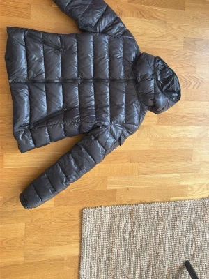 Svart dunjacka från Moncler med huva - Snygg svart dunjacka från Moncler med glansig finish och fast huva. Jackan har två snedställda dragkedjefickor framtill och ett Moncler-märke på ärmen. Perfekt för kalla vinterdagar tack vare det värmande dunet och den klassiska quiltade designen.