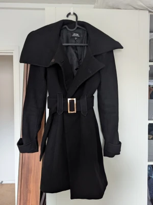 Svart trenchcoat  - Stilren svart kappa  med bred krage, knytskärp i midjan och dekorativt spänne. Jackan har långa ärmar med knappdetalj vid ärmslut. Jackan saknar tyvärr en knapp (se sista bilden), annars är den i väldigt bra skick.