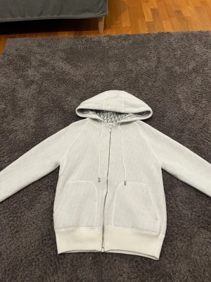 Vit hoodie med dragkedja från Zara - Snygg vit hoodie från Zara med dragkedja framtill och två stora fickor. Huvan har dragsko och insidan är mönstrad med Zara-logga. Ribbstickade muddar vid ärmslut och nederkant. Perfekt för en clean och lyxig streetstyle.