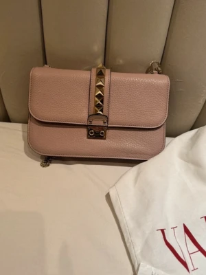 Valentino väska - Säljer min super snygga ljusrosabeige väska från Valentino i äkta läder. Väskan har guldfärgade metalldetaljer, kedjerem och dekorativa nitar framtill. I modellen Glam lock, nypris 20 000 kr🥰 Pris går att diskutera🤗