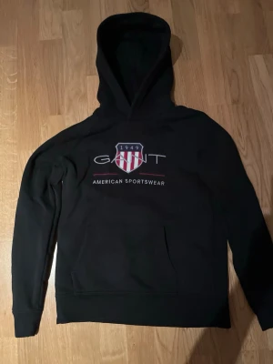  En Svart hoodie från Gant med tryck för en 12-13 åring/xs - Svart hoodie från Gant med klassiskt tryck framtill och broderad logga med amerikansk sköld. Hoodien har känguruficka och dragsko i huvan. Perfekt för en sportig och avslappnad stil.
