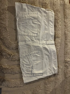 Low waist mini skirt (vit) från Armani Jeans - Snygg vit low waist mini kjol från Armani Jeans (vintage)  med stora fickor fram och bak samt guldfärgad knappdetalj. Kjolen har en rak passform och är tillverkad i ett mjukt bomullstyg. Perfekt för dig som gillar stilrena och klassiska plagg.