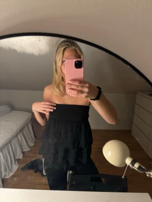 Svart bandeau tubtopp med volang/ kjol - Säljer en svart bandeau tubtopp med volangdetaljer. Toppen är axelbandslös och har en smockad överdel som sitter tight över bröstet. Perfekt för dig som vill ha en trendig och luftig topp till sommaren.