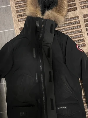 Canada Goose jacka - Säljer en svart parkas från Canada Goose med avtagbar huva och äkta pälsdetalj. Jackan har två stora framfickor, dragkedja med kardborrestängning och klassisk Canada Goose-logga på ärmen. Perfekt för kalla vinterdagar och riktigt snygg streetstil.