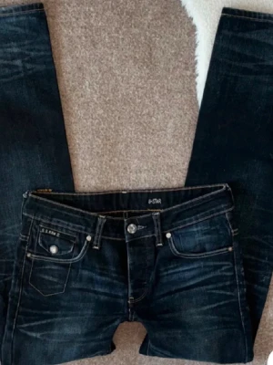 Mörkblå jeans från G-Star RAW - Säljer ett par mörkblå jeans från G-Star RAW med snygga slitningar och kontrastsömmar. Jeansen har raka ben, klassiska fem fickor och coola broderade detaljer på bakfickorna. Materialet är kraftig denim i bomull. Perfekta för en avslappnad och trendig look. Omsydda för att bli mer low rise.