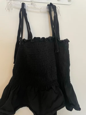 Svart smockad linnetopp från H&M - Säljer en svart linnetopp från H&M med smockad överdel och tunna axelband som knyts upptill. Toppen har volangkant nedtill. Materialet känns mjukt och lätt.