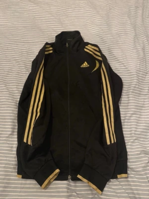 Svart Adidas kofta - Snygg svart track jacket från Adidas med guldfärgade detaljer och tre ränder längs ärmarna. Dragkedja framtill, hög krage och ribbade muddar. Adidas-logga i guld på bröstet.