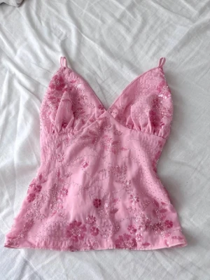 Rosa blommig linnetopp med paljetter - Söt rosa linnetopp med blommigt mönster och paljettdetaljer. Toppen har smala axelband, v-ringning och är figurnära. Perfekt för dig som vill ha en feminin och trendig look.