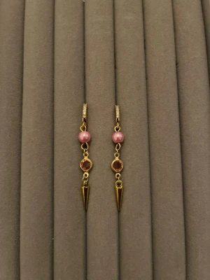 Guldörhängen med rosa pärlor och stenar - Säljer ett par snygga örhängen i guldton med rosa pärlor, rosa stenar och spetsiga hängen. Örhängena har en modern design med små detaljer på ringen och passar perfekt för dig som gillar unika accessoarer. Materialet ser ut att vara metall med pärldetaljer.