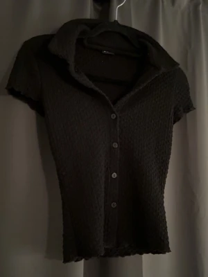 Svart kortärmad skjorttopp - Säljer en svart kortärmad topp med krage och knappar framtill. Tyget är strukturerat och stretchigt vilket ger en snygg passform. Perfekt till jeans eller kjol för en trendig look.