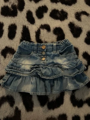 Blå volangkjol i denim med knappar - Supersöt kort jeanskjol i blå denim med volanger och tre dekorativa knappar framtill. Kjolen har fickor fram och resår i midjan för skön passform. Perfekt för dig som gillar en avslappnad men trendig stil.