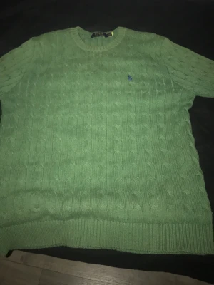 Grön kabelstickad tröja Polo Ralph Lauren - Snygg grön kabelstickad tröja från Polo Ralph Lauren med rund halsringning och klassisk blå logga broderad på bröstet. Tröjan har ribbade muddar vid ärmslut och nederkant. Perfekt för dig som gillar preppy stil och vill ha något bekvämt och stilrent.