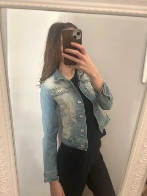 Ljusblå jeansjacka med klassisk passform - Säljer en ljusblå jeansjacka med klassisk krage och silverfärgade knappar. Jackan har två bröstfickor med lock och knapp samt sömdetaljer som ger en snygg look. Perfekt att slänga över en t-shirt eller hoodie för en cool stil.