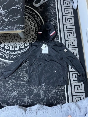 Svart Moncler vindjacka med huva - Snygg svart vindjacka från Moncler med huva och dragkedja. Jackan har vita och röda detaljer på huvan samt Moncler-logga på ärmen. Tillverkad i lätt, glansigt material som står emot vind. Perfekt för dig som vill ha en stilren och exklusiv look.