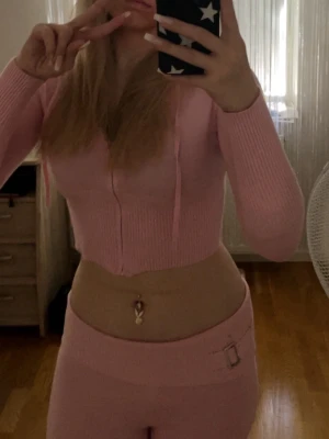 Rosa catania set - Mysigt rosa loungewear-set med croppad hoodie och matchande byxor. Setet är ribbstickat och har en snygg detalj med spänne på byxan. Perfekt för chill dagar hemma. Materialet ser mjukt och stretchigt ut.