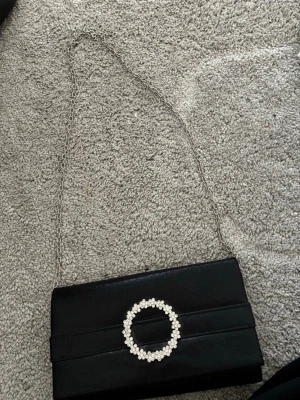 Svart kuvertväska med strassdetalj - Stilren svart kuvertväska med glittrande strasscirkel framtill och tunn silverfärgad kedja. Väskan har en slät, elegant yta och klassisk rektangulär form. Perfekt accessoar för att lyfta din outfit.