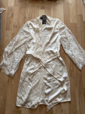 Vit spetsmorgonrock från Hunkemöller - Elegant vit morgonrock från Hunkemöller, dekorerad med vackra spetsdetaljer på ärmar och framstycke. Modellen har trekvartsärmar och ett matchande midjeband i satin. Perfekt för en lyxig och bekväm känsla hemma. Den är helt ny med prislapp och har aldrig använts. Ordinariepris: 549kr