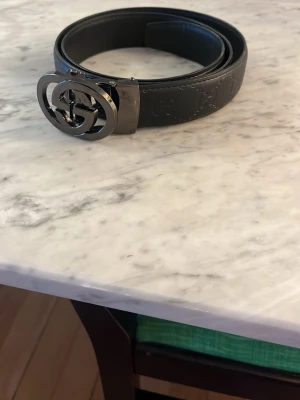 Svart skärp med GG-logo från Gucci - Säljer ett svart skärp från Gucci med den klassiska GG-logon i silver på spännet. Skärpet är gjort av skinn och har präglade GG-mönster längs hela bandet. Perfekt för att lyfta din outfit med en lyxig touch.