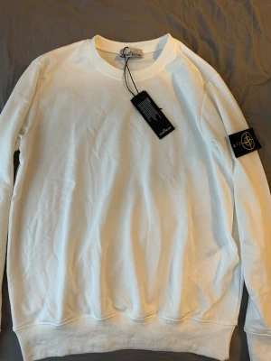 Vit sweatshirt från Stone Island - Säljer en clean vit sweatshirt från Stone Island med klassisk rund hals och ribbade muddar. Tröjan har den ikoniska svarta patchen med gul och grön kompass på vänster ärm. Perfekt för dig som gillar streetwear och stilrena plagg.