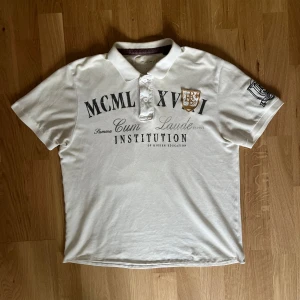 Vit pikétröja - Vit pikétröja från Institution med svart texttryck och broderat emblem på bröstet. Klassisk krage med knappar och korta ärmar. Tröjan har en stilren och sportig look, perfekt för en avslappnad men ändå snygg outfit.