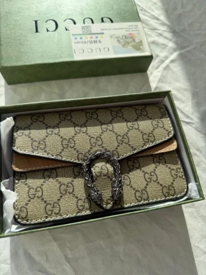 Gucci beige monogram handväska - Säljer en beige handväska från Gucci med klassiskt GG-monogram över hela väskan. Väskan har en silverfärgad dekorativ spänne framtill och detaljer i brunt läder. Kommer i originalask. Perfekt för dig som gillar lyxiga accessoarer och tidlös design.