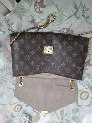 Louis Vuitton monogram axelremsväska - Snygg handväska från Louis Vuitton i klassiskt brunt monogrammönster och beige läderdetaljer. Väskan har guldfärgad kedjerem och elegant guldfärgat lås framtill. Perfekt för dig som vill ha en ikonisk och trendig accessoar.