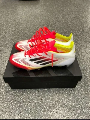 Adidas vita och röda fotbollsskor - Snygga fotbollsskor från Adidas i vitt med röda och gula detaljer samt svarta ränder på sidan. Skorna har en låg profil, tunna syntetmaterial och röda snören. Perfekta för snabba spelare som vill sticka ut på planen.