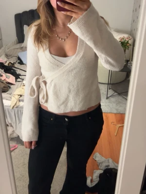 Vit omlottkofta med knytband - Snygg vit omlottkofta med knytband i sidan och långa ärmar. Koftan har en djup v-ringning och är stickad i ett mjukt material. Perfekt för lager-på-lager och ger en chill men ändå stilren look.