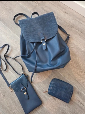 Mörkblå ryggsäck med croco-detaljer - Snygg mörkblå ryggsäck i fejkläder med croco-mönstrat lock och guldfärgade detaljer. Setet inkluderar även en matchande mobilväska och plånbok i samma stil.