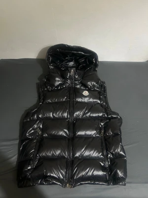 Svart dunväst från Moncler - Snygg svart dunväst från Moncler med glansig finish och huva. Västen har två dragkedjefickor, Moncler-logga på bröstet och quiltad design. Perfekt för lager-på-lager och streetwear-stil.