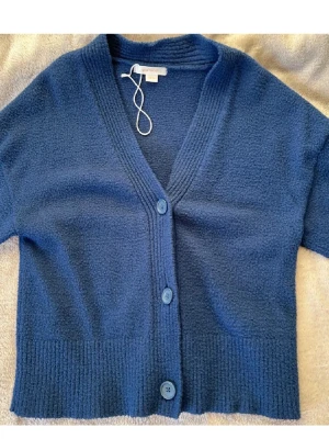 Blå kofta från Gina Tricot - Mysig blå kofta från Gina Tricot med tre knappar framtill och djup V-ringning. Koftan har ribbade muddar och är tillverkad i ett mjukt, stickat material. Perfekt för lager på lager och enkel att matcha med olika outfits.