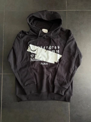 Svart hoodie Maison Margiela Paris - Svart hoodie från Maison Margiela Paris med vit tryckt grafik och siffror på bröstet. Klassisk huva med dragsko och ribbade muddar. Oversized passform och mjukt material, perfekt för en trendig streetwear-look.
