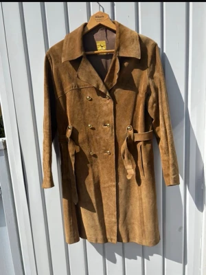Brun mocka trenchcoat med bälte - Snygg brun trenchcoat i mocka från Alberts med dubbelknäppning och matchande bälte i midjan. Jackan har två fickor framtill. Perfekt för dig som gillar retrostil och vill ha en unik jacka i garderoben.